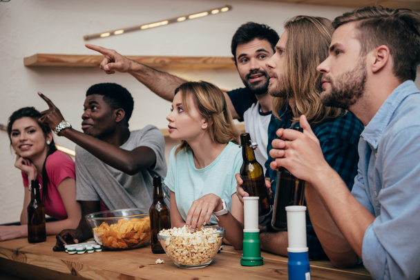kaksi miestä osoittaa käsin ystäviä istuu baarissa tuuletin sarvet, popcorn, sirut ja olut pullot aikana katsella jalkapallo-ottelun
 - Valokuva, kuva