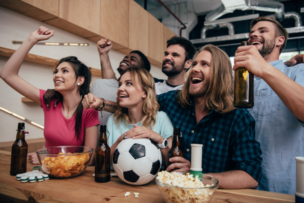 heureux groupe multiethnique d'amis célébrer, faire des gestes oui et regarder match de football au bar avec des chips, bière et ballon de football
  - Photo, image