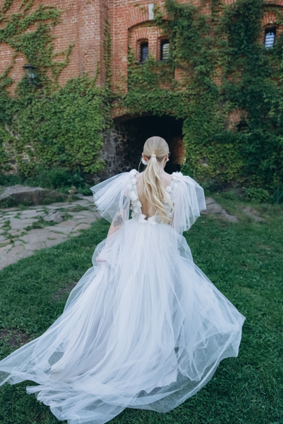 Rückansicht der jungen Braut im wunderschönen Brautkleid vor einem antiken Gebäude, das mit Weinreben bedeckt ist - Foto, Bild