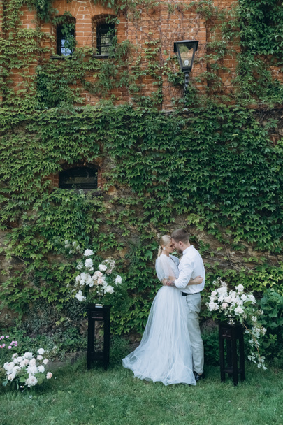Braut und Bräutigam umarmen sich vor einem mit Weinreben und grünen Blättern bedeckten Gebäude - Foto, Bild