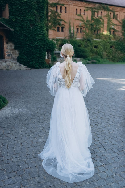 rear view of young bride in beautiful wedding dress in front of ancient building covered with vine - Fotografie, Obrázek