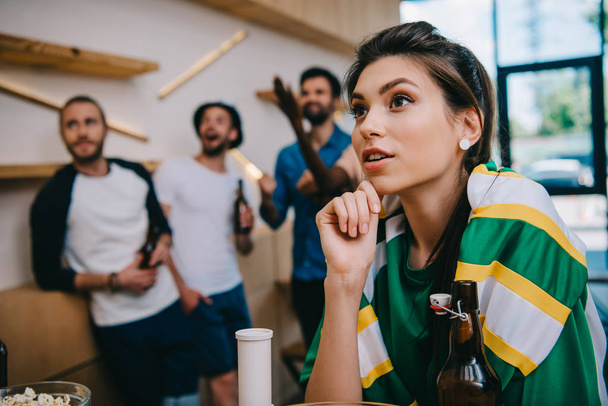 женщина в зеленой футболке с веером и шарфом и ее друзья мужского пола, стоящие позади во время просмотра футбольного матча в баре
  - Фото, изображение