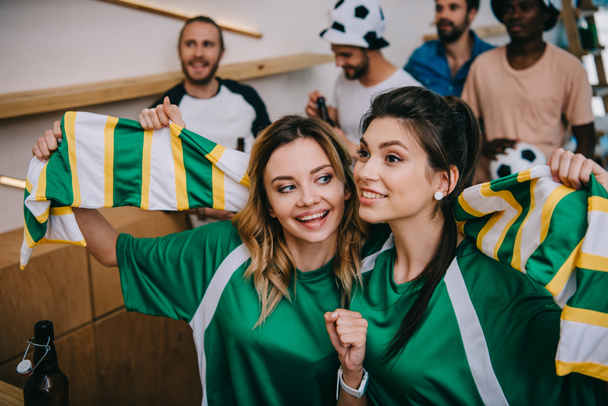 women in green fan t-shirts holding fan scarf and their male friends standing behind during watch of soccer match at bar  - Photo, Image