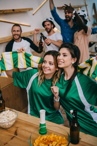 μεγάλη γωνία προβολής των γυναικών στο πράσινο ανεμιστήρα t-shirts κρατώντας ανεμιστήρα κασκόλ και τους αρσενικούς φίλους στέκεται πίσω από, κατά τη διάρκεια ρολόι του αγώνα ποδοσφαίρου στο μπαρ  - Φωτογραφία, εικόνα