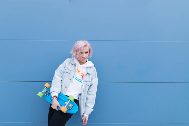 mooi meisje stijl poseren met een rots op de achtergrond van een blauwe muur. Portret van een meisje-blonde die stond op een blauwe achtergrond met een skate in haar armen. Copyspace - Foto, afbeelding