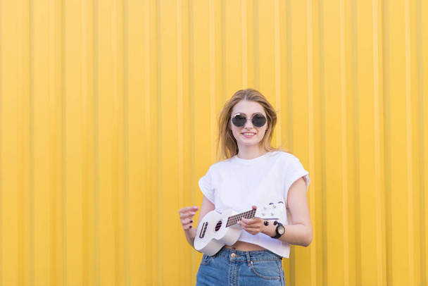 Atraktivní, stylová holka stojí na pozadí žluté zdi s ukulele ve svých rukou a se dívá na kameru. Hudební koncept - Fotografie, Obrázek