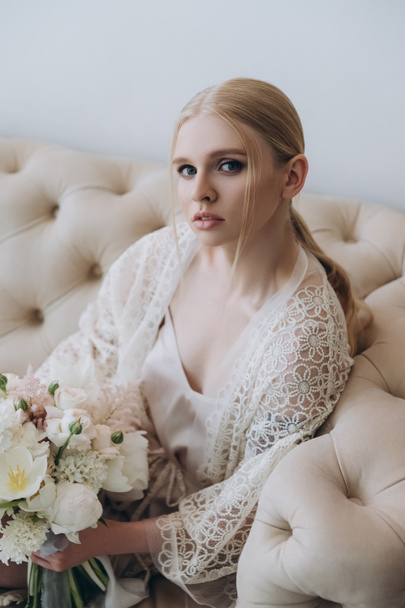 attractive young woman sitting on couch with bouquet looking at camera - Fotografie, Obrázek