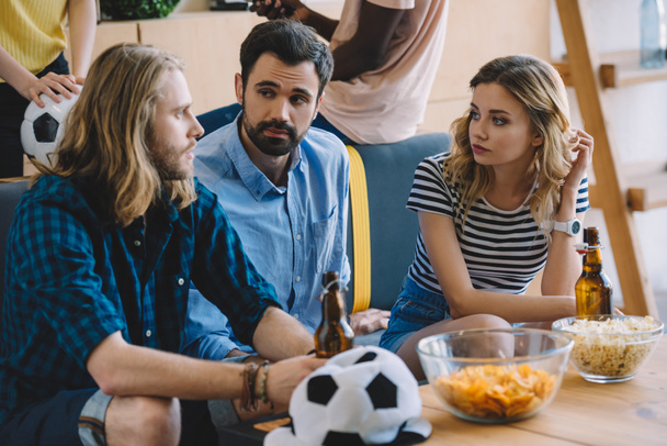 jovens fãs de futebol sentados no sofá com garrafas de cerveja e conversando uns com os outros perto da mesa com batatas fritas e pipocas
  - Foto, Imagem