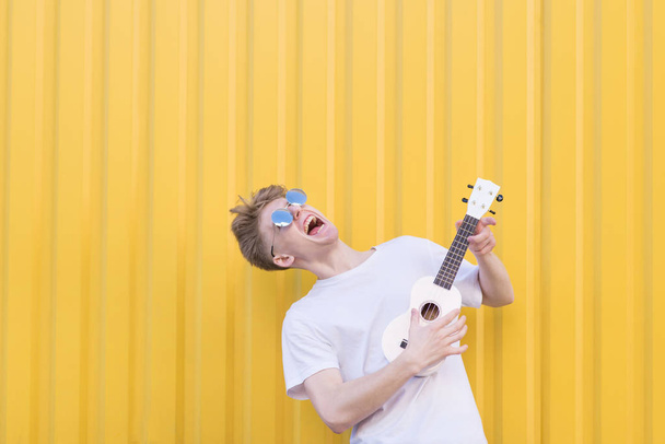Сумасшедший молодой человек играет на укулеле на желтом фоне. Экспрессивный музыкант, играющий на гитаре. Музыкальная концепция
. - Фото, изображение
