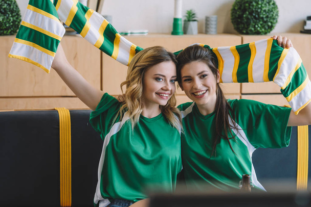 smiling female football fans in green t-shirts and scarf celebrating during watch of soccer match at home - Zdjęcie, obraz