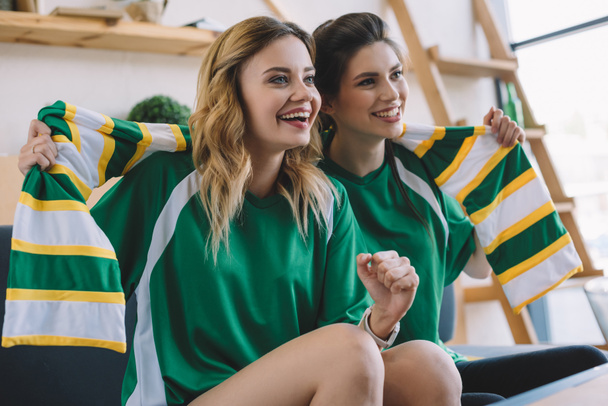 happy female football fans in green t-shirts and scarf gesturing by hands during watch of soccer match at home - Фото, изображение