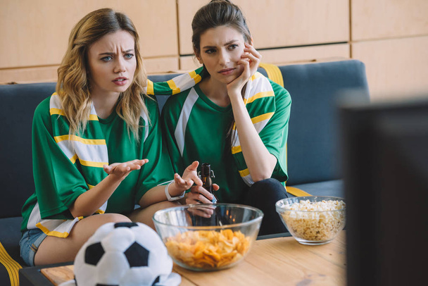 twee vrouwelijke voetbalfans in groene t-shirts verstoord en sjaal gebaren van handen tijdens horloge van voetbalwedstrijd thuis - Foto, afbeelding