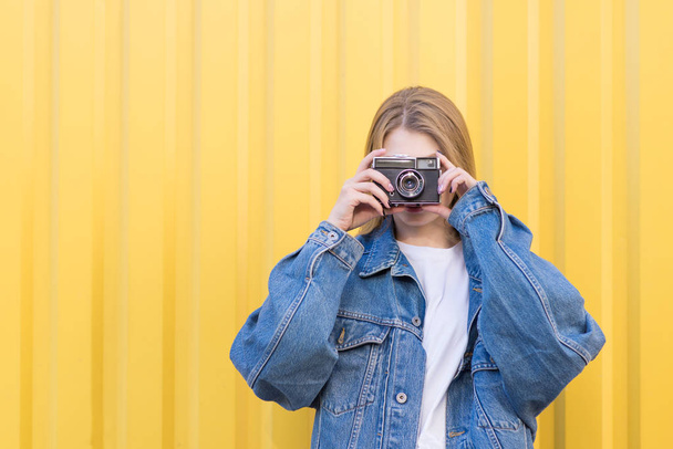 Hipsters meisje is op een gele achtergrond en neemt een foto van een oude filmcamera. Het meisje in een jeans jasje is gefotografeerd op een film. - Foto, afbeelding