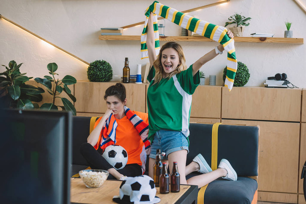 excited woman in green fan t-shirt celebrating victory and holding scarf over head while her upset female friend in orange t-shirt sitting near on sofa during watch of soccer match at home - Фото, изображение