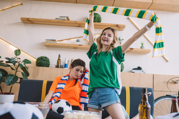 joven mujer sonriente en verde ventilador camiseta celebrando la victoria y sosteniendo bufanda sobre la cabeza mientras que su amiga molesta en naranja camiseta sentada detrás durante el reloj del partido de fútbol en casa
 - Foto, imagen