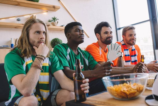 group of multicultural male friends in green and orange fan t-shirts watching soccer match near table with chips and popcorn at home - Foto, afbeelding