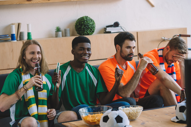 χαμογελώντας τους οπαδούς του ποδοσφαίρου σε πράσινο μπλουζάκια για τον εορτασμό ενώ αναστατωμένος φίλους τους πορτοκαλί μπλουζάκια που κάθονται κοντά στον καναπέ κατά τη διάρκεια ρολόι του ποδοσφαίρου ταιριάζει στο σπίτι - Φωτογραφία, εικόνα