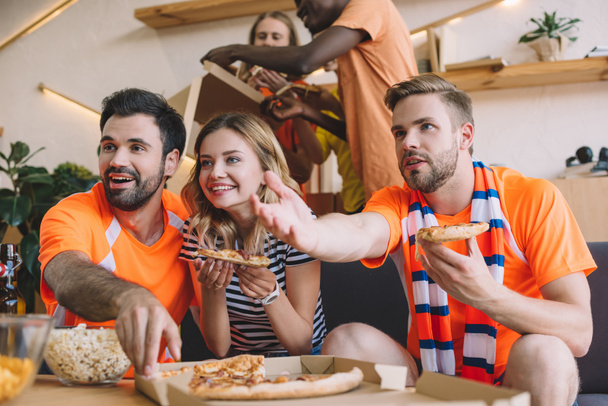 Nuori mies osoittaa käsin ja hänen ystävänsä syövät pizzaa ja katsovat jalkapallo-ottelua kotona
 - Valokuva, kuva