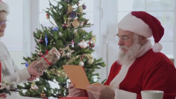 Santa Claus in a red costume sitting on the table, read a letter. Suddenly small boy came and give to grandfather a present. Old man wanted to kiss a kid to thank him, but boy did not want. He keep - Кадри, відео