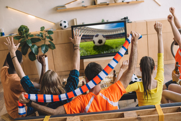 молодые футбольные болельщики празднуют и делают "да" жесты, сидя на диване во время просмотра футбольного матча дома
 - Фото, изображение