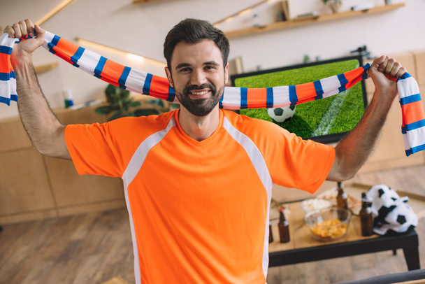 portrait de jeune homme en t-shirt orange fan tenant foulard rayé et célébrant la victoire dans le match de football à la maison
  - Photo, image