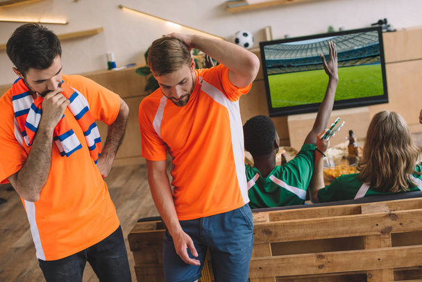 deux fans de football masculin contrariés en t-shirts orange et leurs amis en t-shirts verts gesticulant et célébrant la victoire pendant la montre du match de football à la maison
  - Photo, image