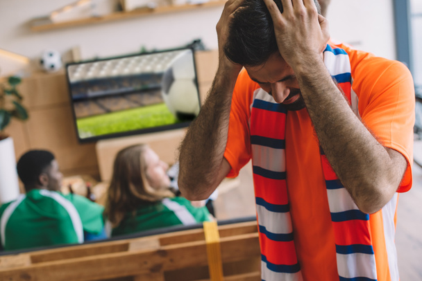 upset young man in orange t-shirt and scarf holding hands on head while his friends watching soccer match on tv screen at home  - Фото, изображение