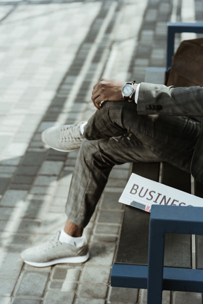Cropped view of African american businessman wearing suit sitting on bench with newspaper - Photo, Image