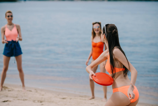 young female friends in swimwear playing with flying disc on sandy beach - Φωτογραφία, εικόνα
