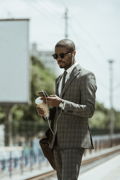 Young african american businessman listening to music and holding coffee cup on train station - Photo, Image