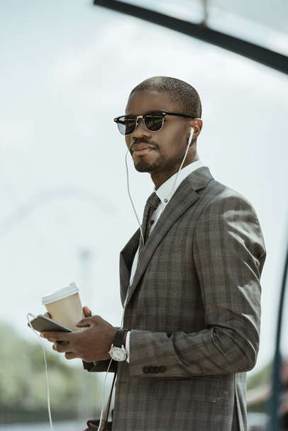 音楽を聴くと、公共交通機関の駅でコーヒー カップを保持している若いアフリカ系アメリカ人のビジネスマン - 写真・画像