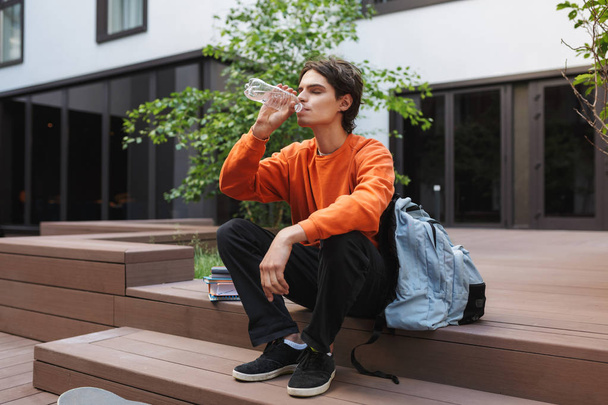 Young man sitting and drinking water with big backpack near in courtyard of university  - Fotoğraf, Görsel