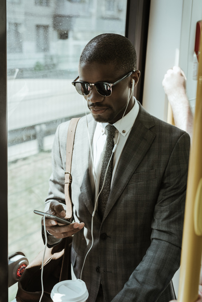 African american businessman wearing suit in earphones using smartphone while taking train - Foto, Imagem
