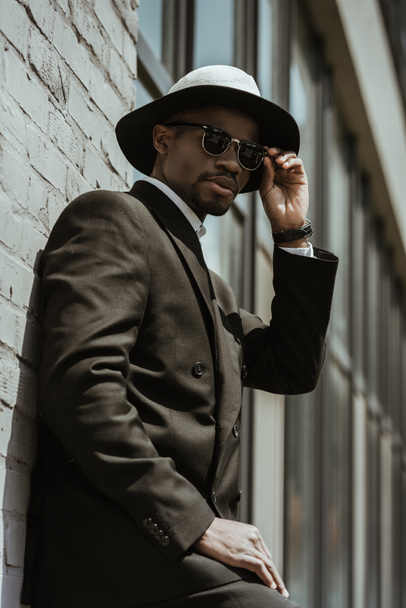 Fashionable african american businessman wearing fedora hat and sunglasses posing by building wall  - 写真・画像