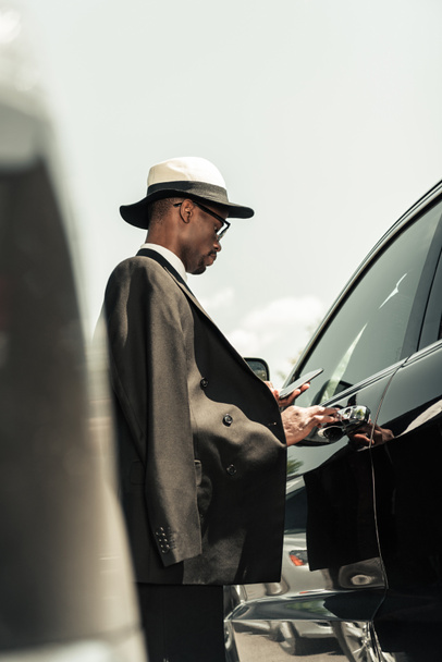 Elegante hombre con camisa blanca y chaqueta que sostiene el teléfono inteligente y abre la puerta del coche
 - Foto, imagen