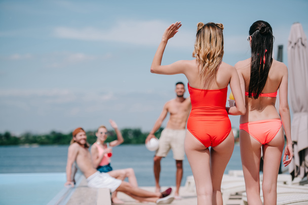 back view of girls in swimwear walking on beach and waving hand to friends at poolside - Fotoğraf, Görsel