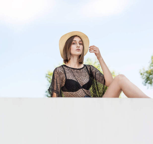 Hermosa chica joven delgada en un sombrero de paja se sienta en una cerca blanca
 - Foto, Imagen