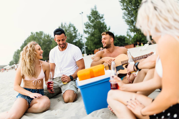 Group of attractive young people enjoying at beach, drinking and playing guitar. - Foto, Imagem