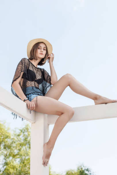Hermosa chica joven delgada en un sombrero de paja se sienta en una cerca blanca
 - Foto, imagen