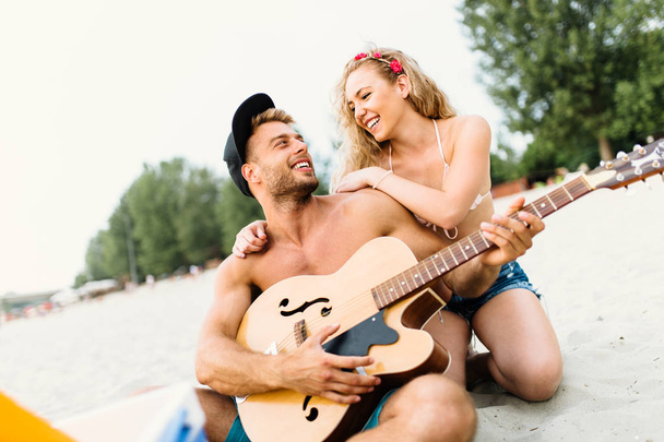 Couple of attractive young people enjoying at beach, drinking and playing guitar. - Fotografie, Obrázek