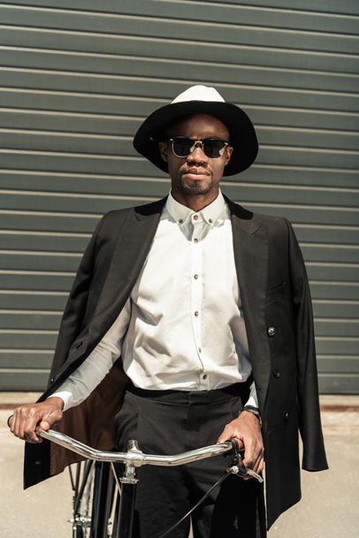 Fashionable african american businessman wearing fedora hat and holding bike - Photo, Image