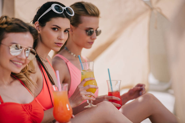 attractive girlfriends in swimwear and sunglasses drinking cocktails in bungalow on beach  - Foto, Imagem