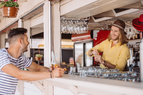 glimlachend barman en client met bierfles kijken elkaar in de strandbar - Foto, afbeelding