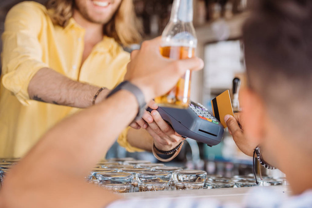 tiro recortado de jovem segurando garrafa de cerveja e pagar com cartão de crédito no bar da praia
 - Foto, Imagem