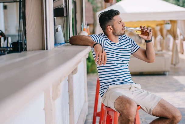 young man drinking beer from glass bottle while sitting at beach bar  - Foto, Imagen