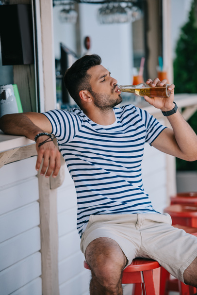 joven guapo bebiendo cerveza de botella de vidrio mientras está sentado en el bar de la playa
  - Foto, Imagen