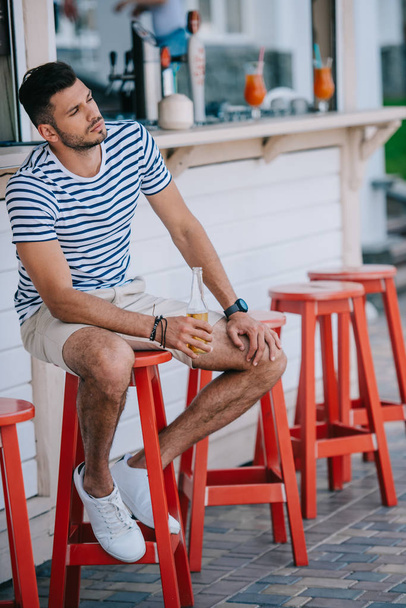 pohledný mladý muž držel sklenici piva zatímco sedí v plážovém baru - Fotografie, Obrázek