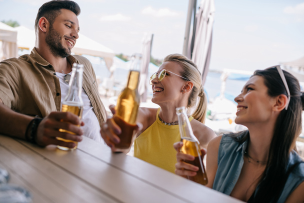 šťastný mladý muž při pohledu na krásnou usmívající se dívky a popíjeli pivo společně v plážovém baru - Fotografie, Obrázek