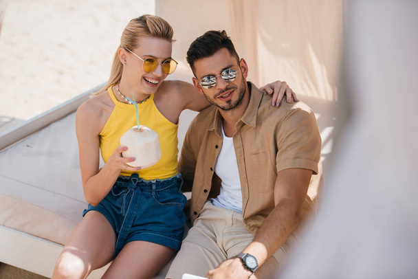 happy young couple in sunglasses drinking summer cocktail while sitting together in bungalow at beach - 写真・画像