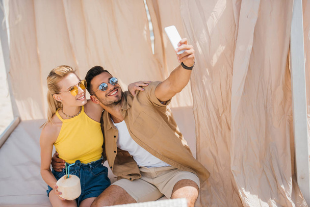 happy young couple in sunglasses taking selfie with smartphone in bungalow  - Photo, Image